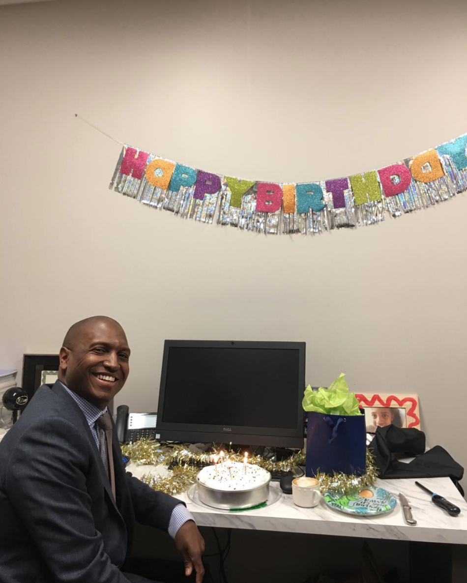 Scott M. Perryman, MD, FACS, FASMBS smiling at his desk with a birthday cake and banner from the office team